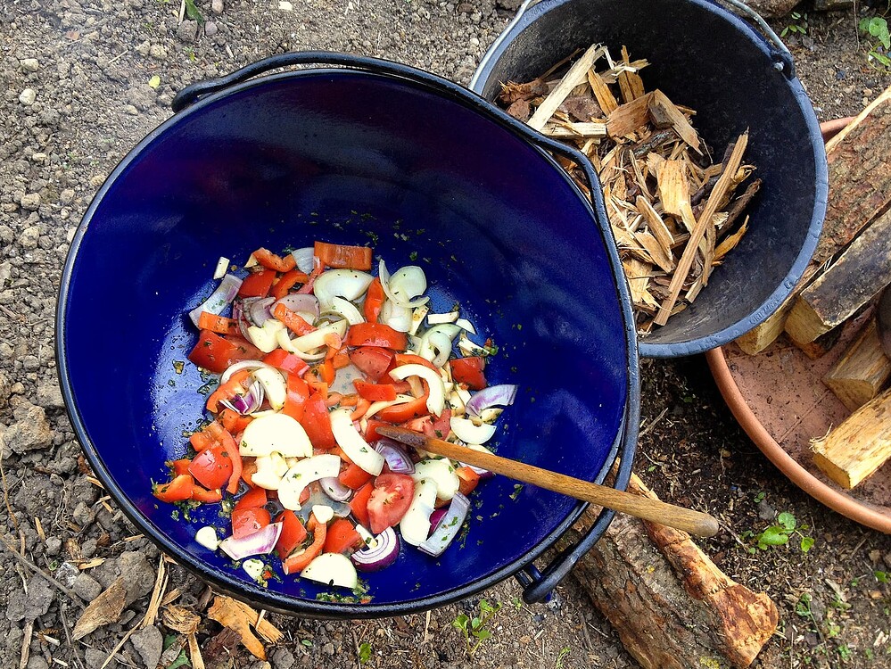 Ein blauer Kessel über einem offenen Feuer, in dem Gemüse wie Paprika und Zwiebeln geschmort wird, daneben ein Eimer mit Brennholz.