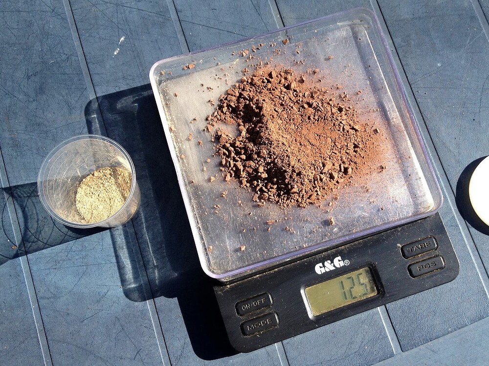 Eine digitale Küchenwaage mit braunem Pulver (Urgesteinsmehl) und daneben ein kleiner Becher mit hellem Pulver (Dolomitkalk).