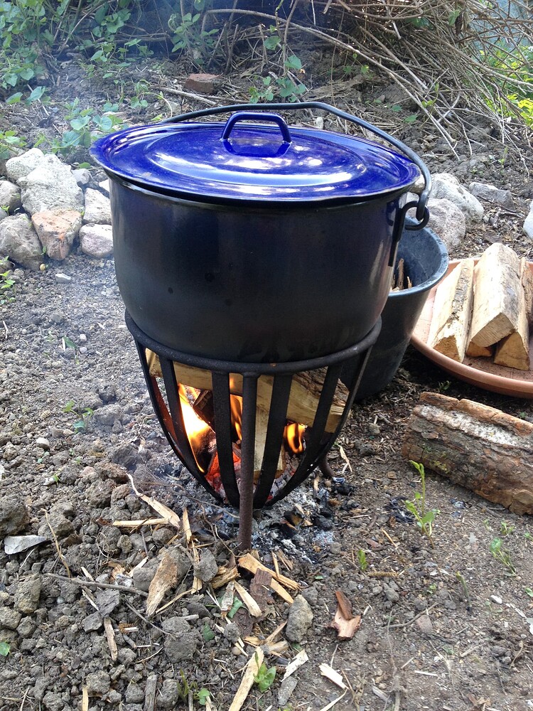 Ein blauer Emaille-Topf steht auf einem Metallgestell über einem offenen Feuer im Freien.