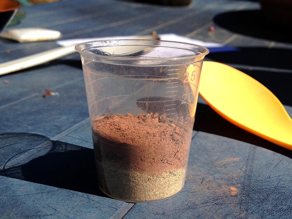 Ein Messbecher mit zwei Schichten Pulver, Dolomitkalk und Urgesteinsmehl, auf einem blauen Tisch.