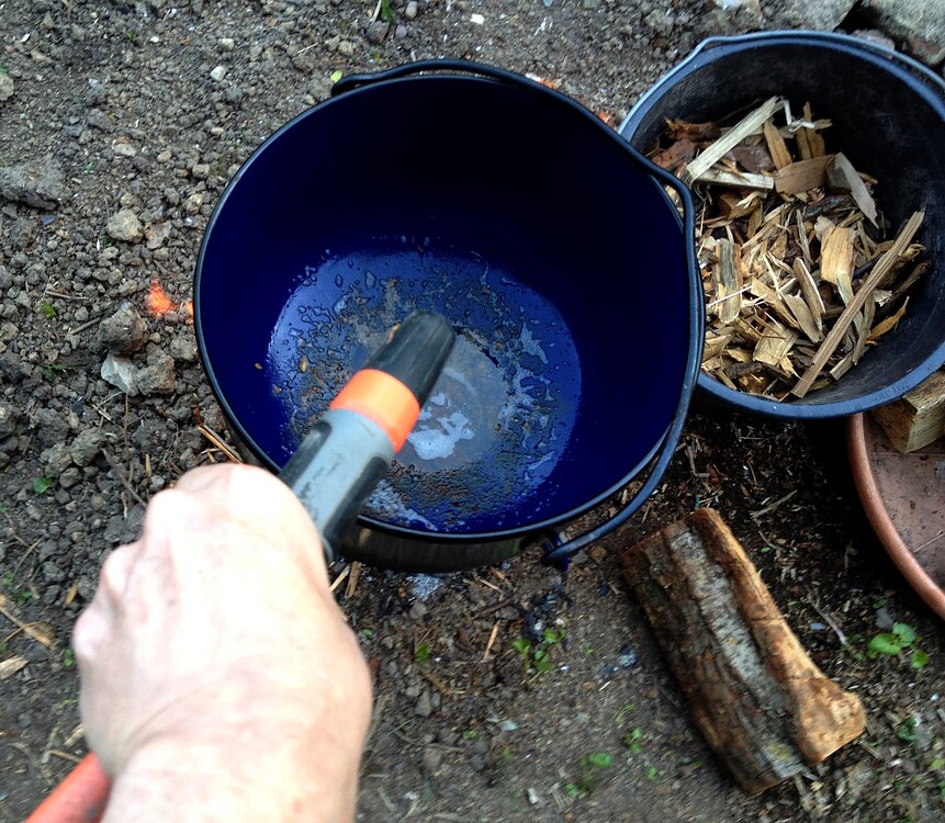 Ein blauer gusseiserner Topf über einem Feuer, in dem gerade gekocht wird, daneben ein Eimer mit Holz und ein Holzscheit auf dem Boden.