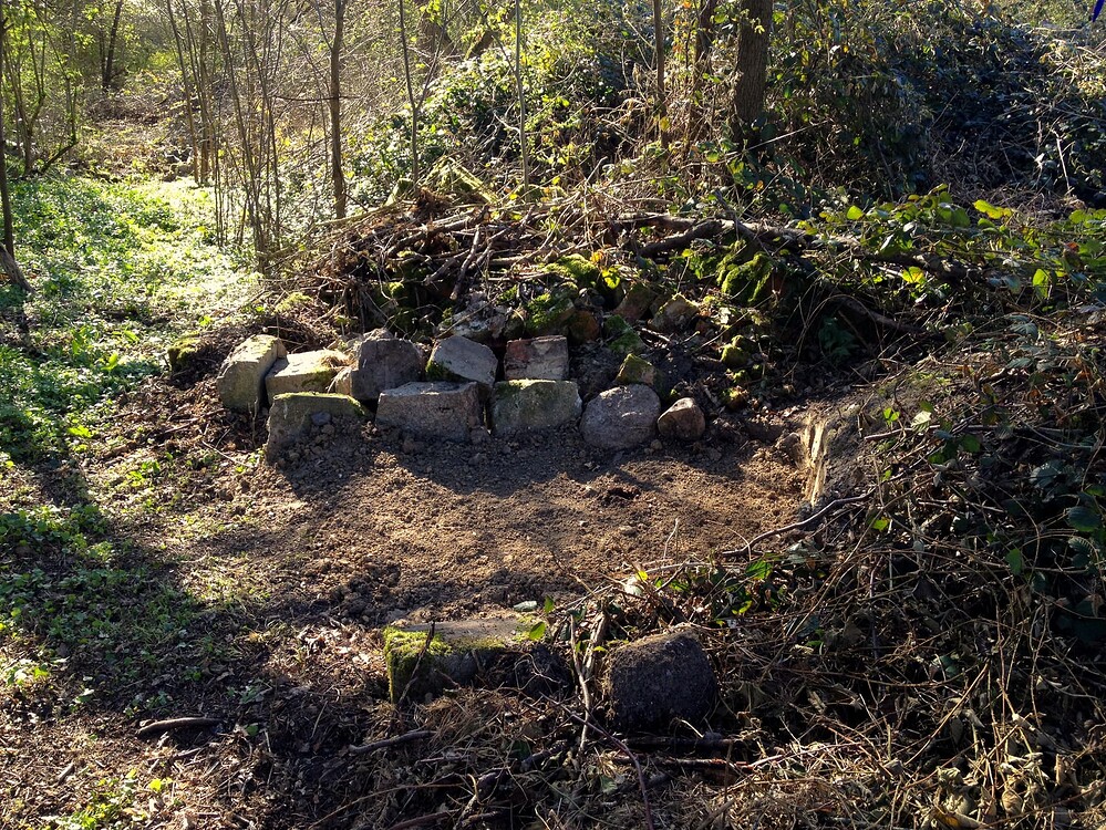 Ein Gartenbereich mit einem neu angelegten Kompostplatz, der mit Steinen eingefasst ist, umgeben von Bäumen und Unterholz.