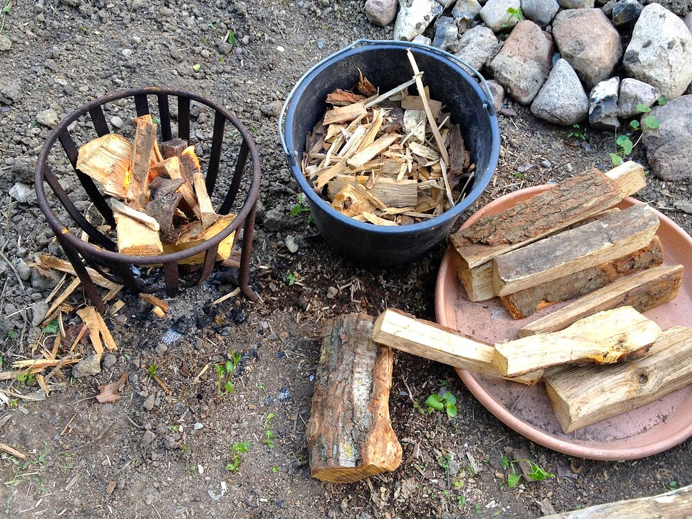 Eine Feuerstelle im Freien mit einem Metallkorb voller brennendem Holz, einem Eimer mit Holzresten und einem Tonteller mit Holzscheiten auf dem Boden.