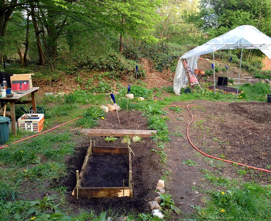 Ein Gartenbereich mit einem hölzernen Hochbeet-Rahmen im Vordergrund, einem kleinen provisorischen Gewächshaus im Hintergrund und verschiedenen Gartenutensilien.